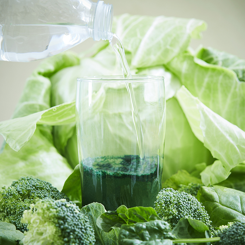 Isotonix Complete Greens, pouring water into partially filled glass of powdered greens, surrounded by leafy greens