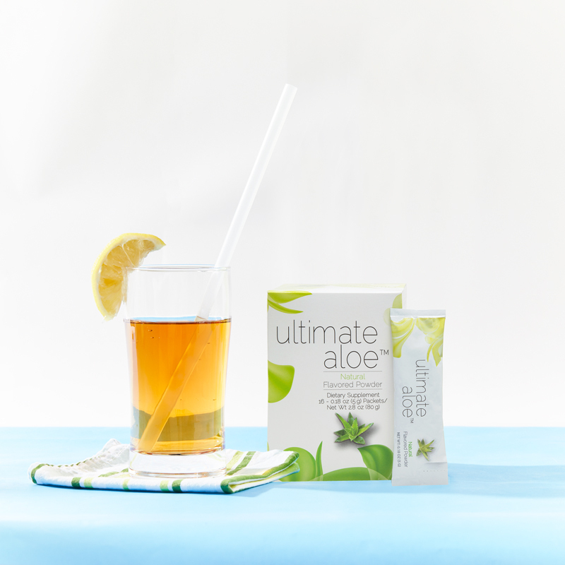 Ultimate Aloe Powder Natural Flavor, with a glass almost filled with liquid, an orange wedge on the brim, and a straw, and packet leaning on Box