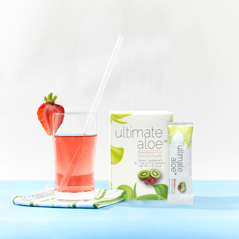 Ultimate Aloe Powder Strawberry Kiwi Flavor, with a glass almost filled with liquid, a strawberry on the brim, and a straw, and packet leaning on Box
