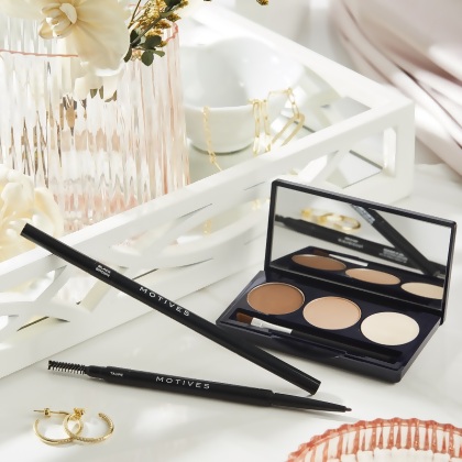 Motives Essential Brow Kit on a vanity, with a vase of orchids and a pair of earrings
