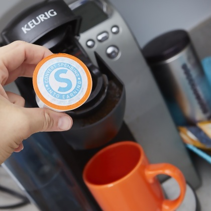 Shopping Annuity Brand Premium Coffee pod shown over coffee maker and an orange cup