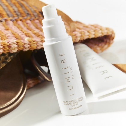 Lumière de Vie Daily Brightening Broad Spectrum SPF 50 pump bottle, next to a straw purse