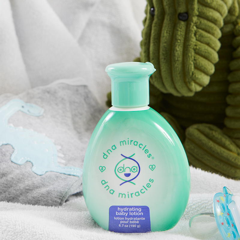 DNA Miracles Natural Hydrating Baby Lotion, on a towel with a pacifier and stuffed toys 