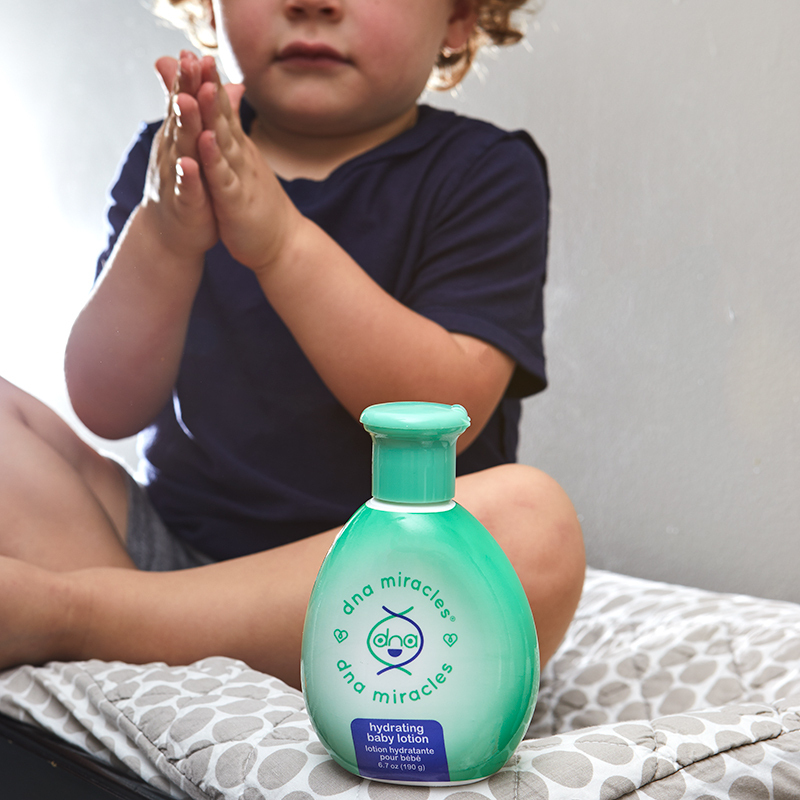 DNA Miracles Natural Hydrating Baby Lotion, with small child sittimg on a towel rubbing product on hands