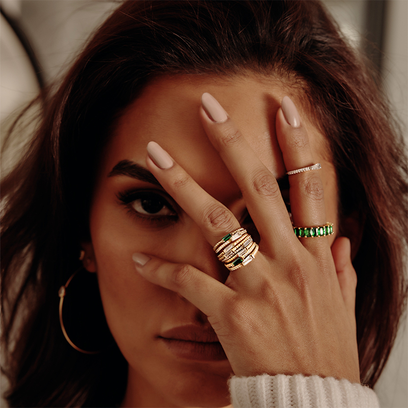Model wearing 8 Layered Adrienne - Spiraled Baguette Rings, stacked, with green stones and clear stones, in gold, with other rings