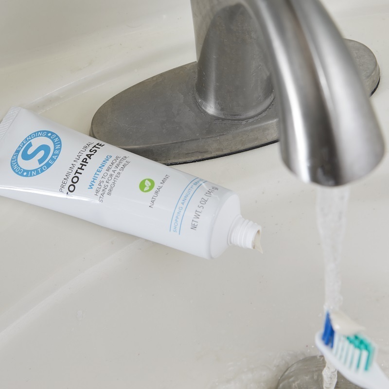 Shopping Annuity Brand Premium Natural Toothpaste on a sink with a toothbrush under running water