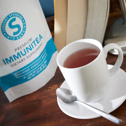 Shopping Annuity Brand Premium ImmuniTea shown with white cup filled with tea-colored liquid, with books in the background